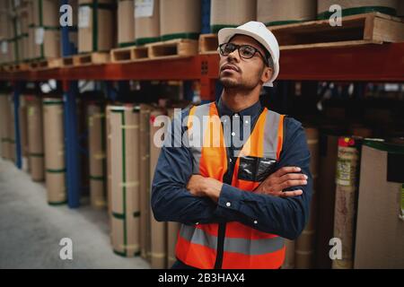 Young warehouse manager standing with arms crossed in factory with white helmet looking away Stock Photo