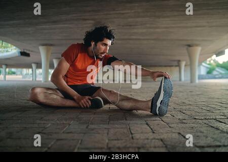 Male athlete enjoying music on earphone warming up and stretching his leg while relaxing under the concrete bridge Stock Photo