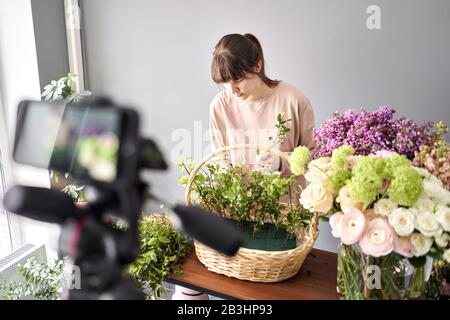 A woman writes a video blog or online course. Flower school concept. Florist woman creates flower arrangement in a wicker basket. Flowers delivery. Stock Photo