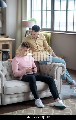 Man in orange shirt sitting on the sofa watching his partner chatting online Stock Photo