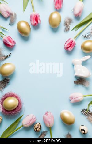 Easter frame of bunny, golden eggs, pink tulips on pastel blue. Top view. Happy Easter holiday. Greeting card. Vertical composition. Stock Photo