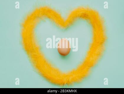 Chicken simple Easter egg in the heart of fluffy yellow feathers on a blue mint clean background. Happy Easter minimalism mockup, flat lay Stock Photo
