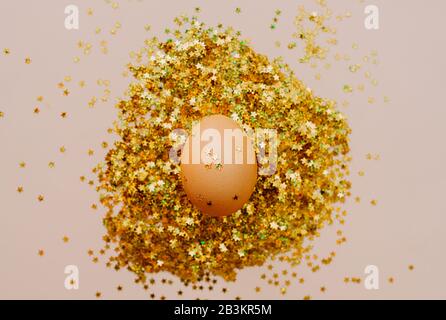 Easter holiday brown egg in a large pile of shiny gold stars on a delicate pink background. Easter layout in art Deco style Stock Photo