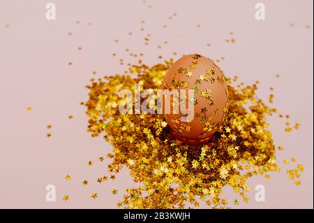 Easter holiday brown egg in a large pile of shiny gold stars on a delicate pink background. Easter layout in art Deco style Stock Photo