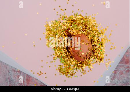 Easter holiday brown egg in a large pile of shiny gold stars on a delicate pink background. Easter layout in art Deco style Stock Photo