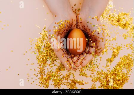 light skin hand in gold spangles in the form of stars firmly hold a Golden Easter egg on a pink background with sequins. Happy Easter flat lay mockup Stock Photo
