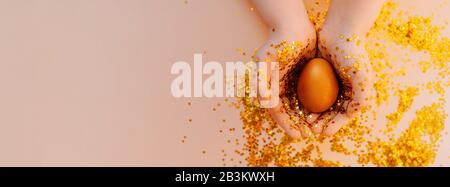 light skin hand in gold spangles in the form of stars firmly hold a Golden Easter egg on a pink background with sequins. Happy Easter flat lay mockup Stock Photo