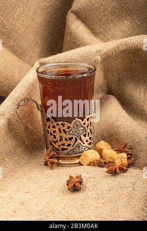 A glass of tea in a vintage Cup holder , star anise and brown sugar on a background of rough homespun fabric. Close up Stock Photo