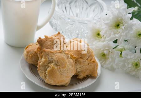 This eclairs are stuffed with vanilla cream. Cakes are situated on the plate against the white colours background: white surface, flowers and the cup. Stock Photo