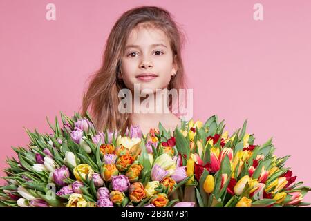 Cute smiling child girl holding bouquet of spring flowers tulips isolated on pink background. Little toddler girl gives a bouquet to mom. Copy space f Stock Photo