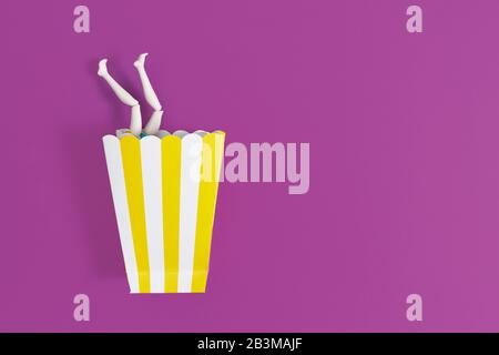 Time for party. Female legs peek out from paper cup for popcorn on a purple background. Copy space for text Stock Photo