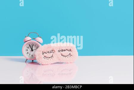 Sleep optimization. Pink fluffy mask for sleeping and a pink alarm clock on a light blue background. Stock Photo