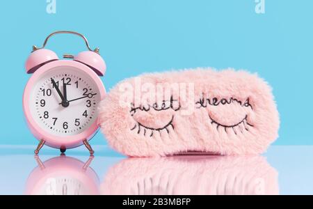 Sleep optimization. Pink fluffy mask for sleeping and a pink alarm clock on a light blue background. Stock Photo