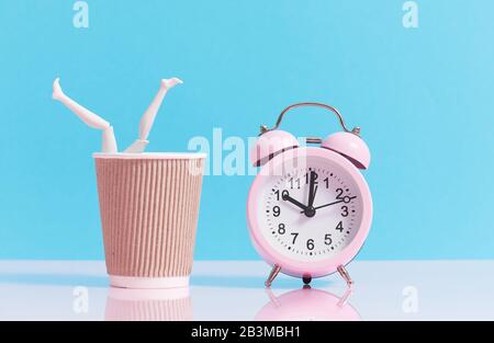 Time to drink some coffee. Female legs peek out from a coffee paper craft cup on a light blue background. Nearby is a pink alarm clock. Stock Photo