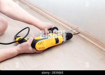 Female hands hold a heavy drill tool and make a hole in the wall for attaching the baseboard. Home repairs. Feminism Stock Photo