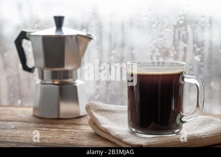 A cup of coffee on the background of geyser coffee machine and window with raindrops. Closeup, selective focus, wooden surface Stock Photo