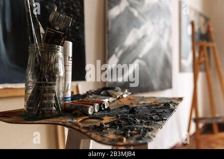 Image of artist workplace with painting tools on table, easel and artworks on wall Stock Photo