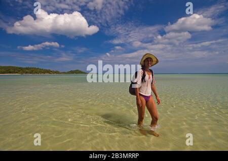 The woman walks on the shallow of lagoon. She has a hat on her head. Stock Photo