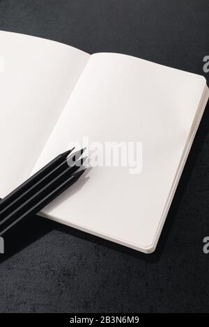 Opened book page with white paper and three black pencils on a black background Stock Photo