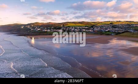 Aerial view of the town of Woolacombe and it's beach at dawn, waves breaking on a beach, the clouds reflected in the wet sand