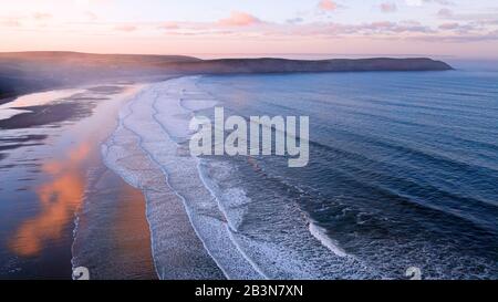 Aerial view of Woolacombe beach at dawn, waves breaking on a beach where the sunlit clouds are reflected in the standing water. Headlands on the horiz Stock Photo