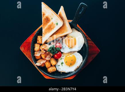 Western brunch style food with toasts, sunny side up egg, tomatoes on a wooden plate and black background. Stock Photo