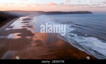 Aerial view of Woolacombe beach at dawn, waves breaking on a beach where the sunlit clouds are reflected in the standing water. Headlands on the horiz Stock Photo