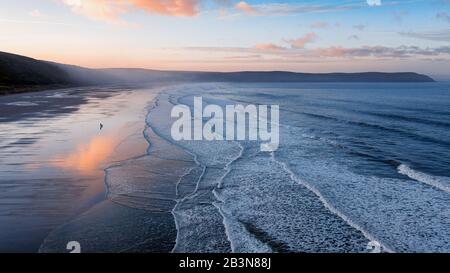 Aerial of Woolacombe beach at dawn, waves breaking on a beach where the sunlit clouds are reflected in the standing water. Distant a man walks his dog Stock Photo