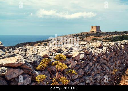 A stone wall leads the eye to a square stone fort on the dingli cliffs on the island of Malta, cloud studded blue sky and the sea in the distance Stock Photo