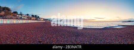 Panorama of the Lyme Regis beach, a row of beach huts and the red shingle pebble beach leading into the distance the sky colored by predawn sunlight Stock Photo