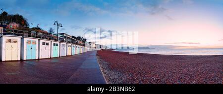 Panorama of Lyme Regis beach, a row of beach huts and the red shingle pebble beach leading into the distance the sky colored by the predawn sunlight Stock Photo