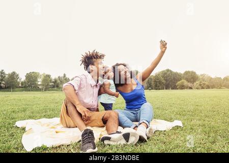 Happy African family doing selfie photo with mobile phone in a public park outdoor - Mother and father having fun with their daughter during a weekend Stock Photo