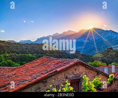 The sun dips behind a mountain peak casting a halo above and rays below. In the foreground the clay tiled roof of a rustic farm building Stock Photo