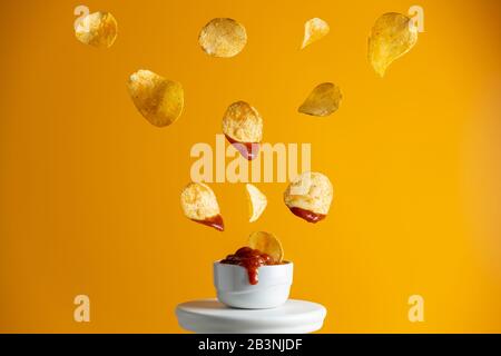 Tasty potato chips falling into blow with tomato sauce, frozen in the air. Yellow food art background. Stock Photo