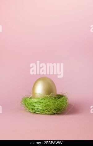 Golden egg in green nest on pink. Easter minimal creative vertical composition with copy space. Close up. Happy Easter holiday. Greeting card. Stock Photo