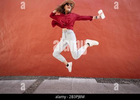 Portrait of an active stylish woman dressed in bright shirt with hat and coffee cup jumping on the red wall background outdoors. Carefree lifestyle, coffee and women's fashion concept