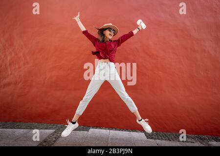 Portrait of an active stylish woman dressed in bright shirt with hat and coffee cup jumping on the red wall background outdoors. Carefree lifestyle, coffee and women's fashion concept Stock Photo