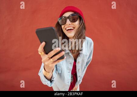Portrait of a young stylish woman feeling happy, chatting on mobile phone on the red wall background outdoors Stock Photo