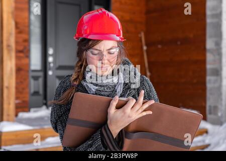 Female Civil engineer holding a clipboard and taking notes outside during cold weather. she wears a red hard hat and Safety Glasses and winter clothes Stock Photo