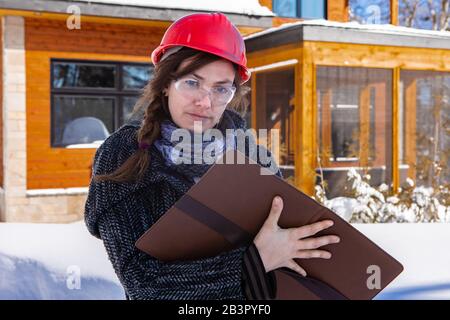 low angle view of a Female Civil engineer standing outdoor wearing a red hard hat and holding a clipboard. the snow around building in the background. Stock Photo