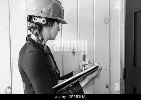 worker woman wears a grey hardhat at work. female construction inspector taking notes on her clipboard during a home inspection, black and white. Stock Photo