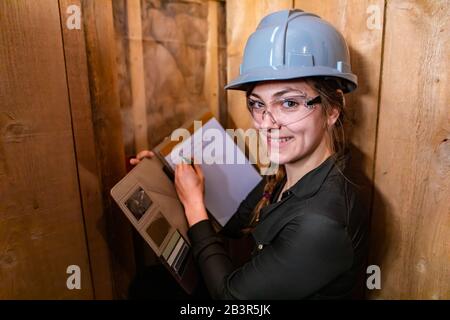 Indoor air quality inspector woman looking for molds or fungi problems on wooden walls and taking notes using her clipboard. she happy and smiling. Stock Photo