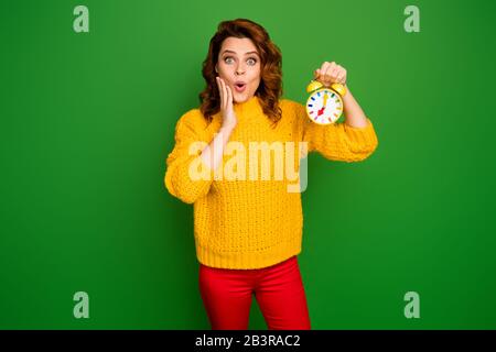 Portrait of astonished woman get up hold clock look time impressed touch hand face scream omg wow wear style trendy pullover isolated over bright Stock Photo