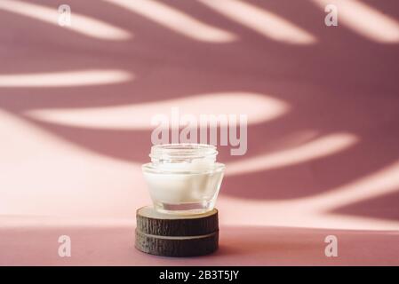 Glass jar with white moisturizing cream on wooden saw cuts on a pink background. Lotion on the background of the tropical shadow of a palm leaf. The c Stock Photo