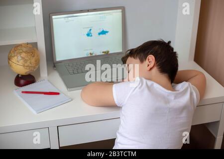 A schoolboy in a white T-shirt fell asleep behind a monitor from a laptop, holding his head in his hands. Home lessons. Exam preparation. hard to lear Stock Photo