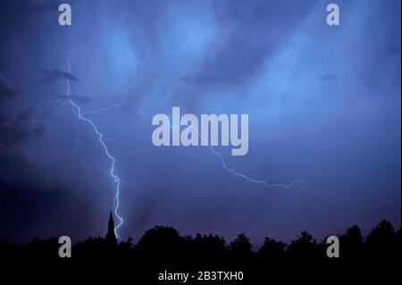 Rain falling and stroke of forked lightning during thunderstorm at night over church tower and trees Stock Photo