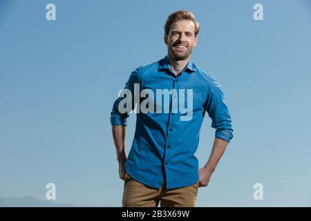 gorgeous casual man wearing blue shirt standing with hands behind him on his back pocket happy outside on blue sky Stock Photo