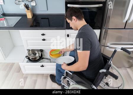 Young Happy Disabled Man Sitting On Wheelchair Arranging Plates In Drawer Stock Photo