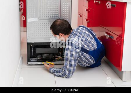 Young Male Serviceman Repairing Broken Refrigerator In Kitchen Stock Photo