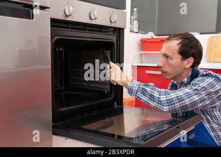 Young Male Technician In Overall Checking Oven In Kitchen Stock Photo